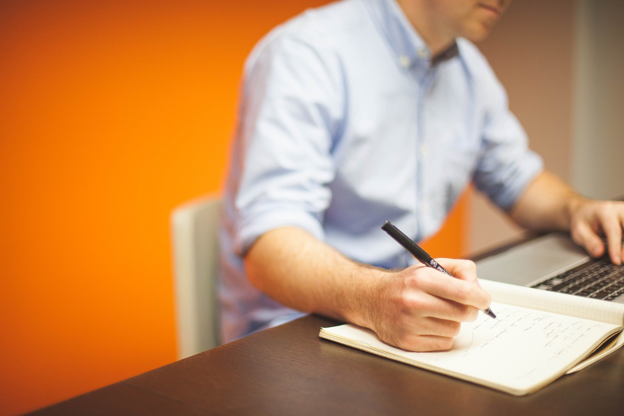 Man using a laptop whilst writing on a notepad, with an orange colour wall as the background