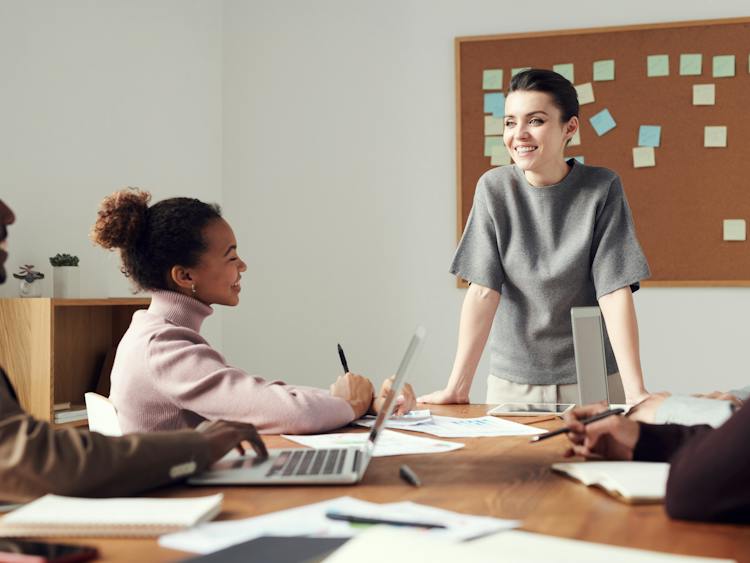 Woman standing at the end of a boardroom table, with paperwork on the table and colleagues sat with computers around the table, collaborating on a project.