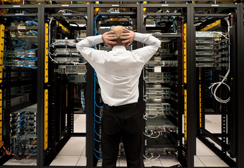 Man standing in front of server racks holding the back of his head in shock / panic