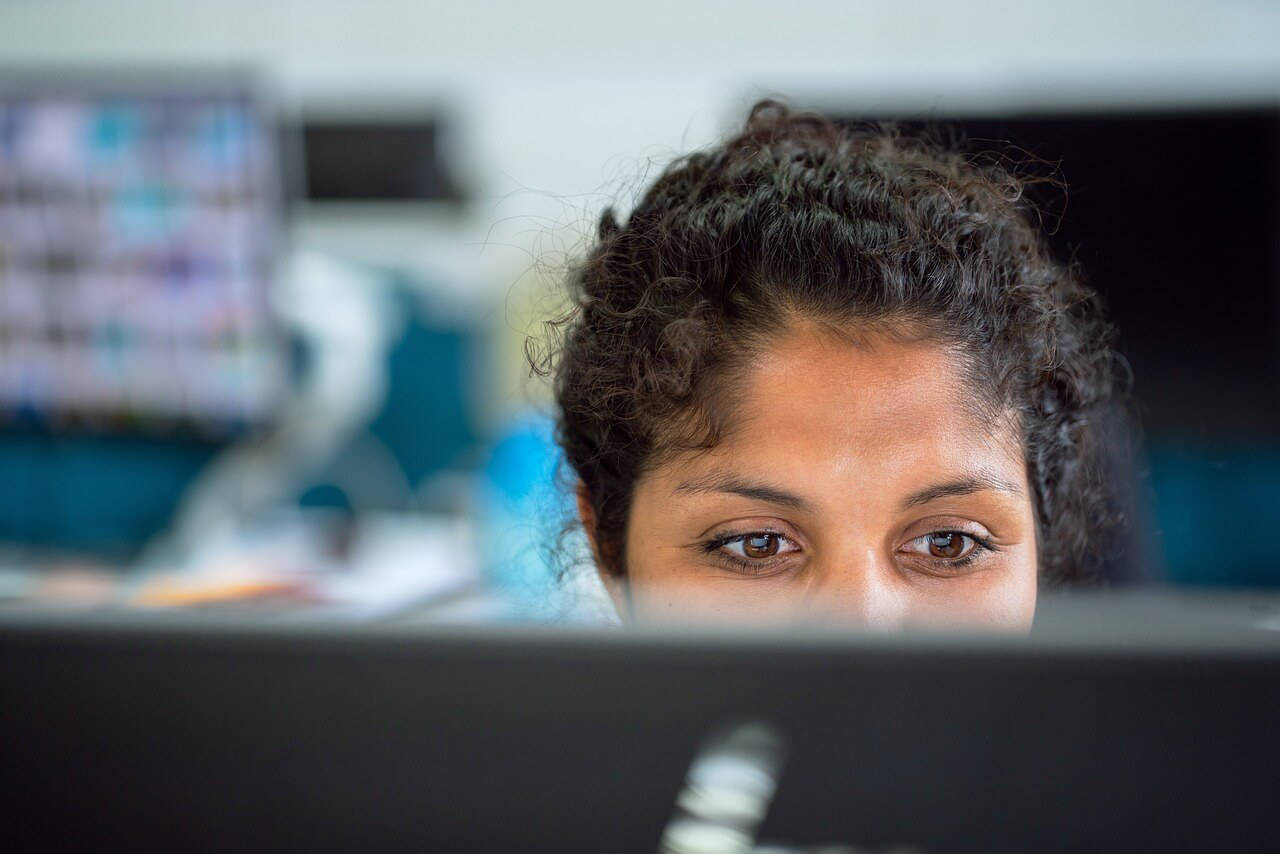 Lady sat at her desk with the top of her head above the computer screen, working on the computer with a blurry background.