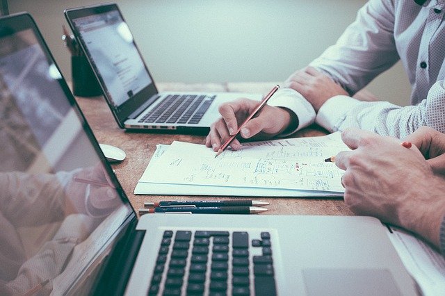 Image depicting a business meeting - two men sat at a table with laptops, a pen and paper, designing a plan